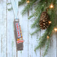 Metal bottle opener with 'Chalet Bar BQ' text hanging on a Christmas tree with pine cones and lights.