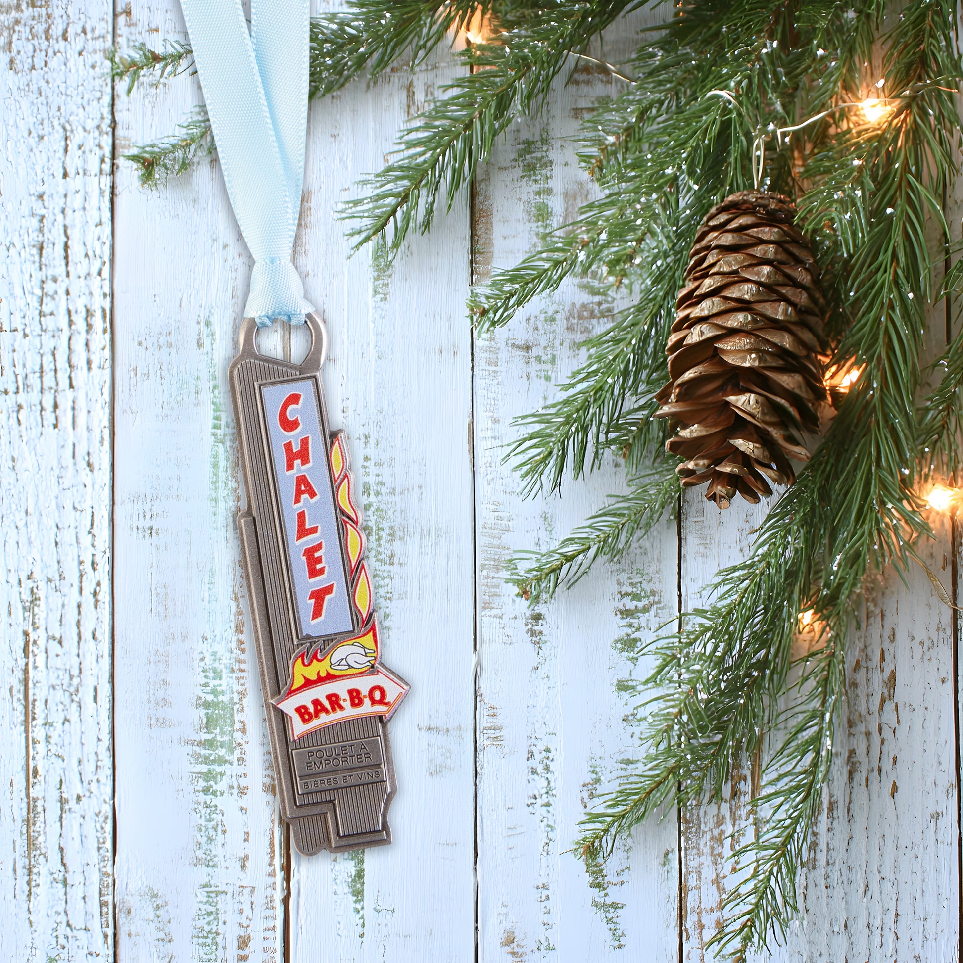 Metal bottle opener with 'Chalet Bar BQ' text hanging on a Christmas tree with pine cones and lights.
