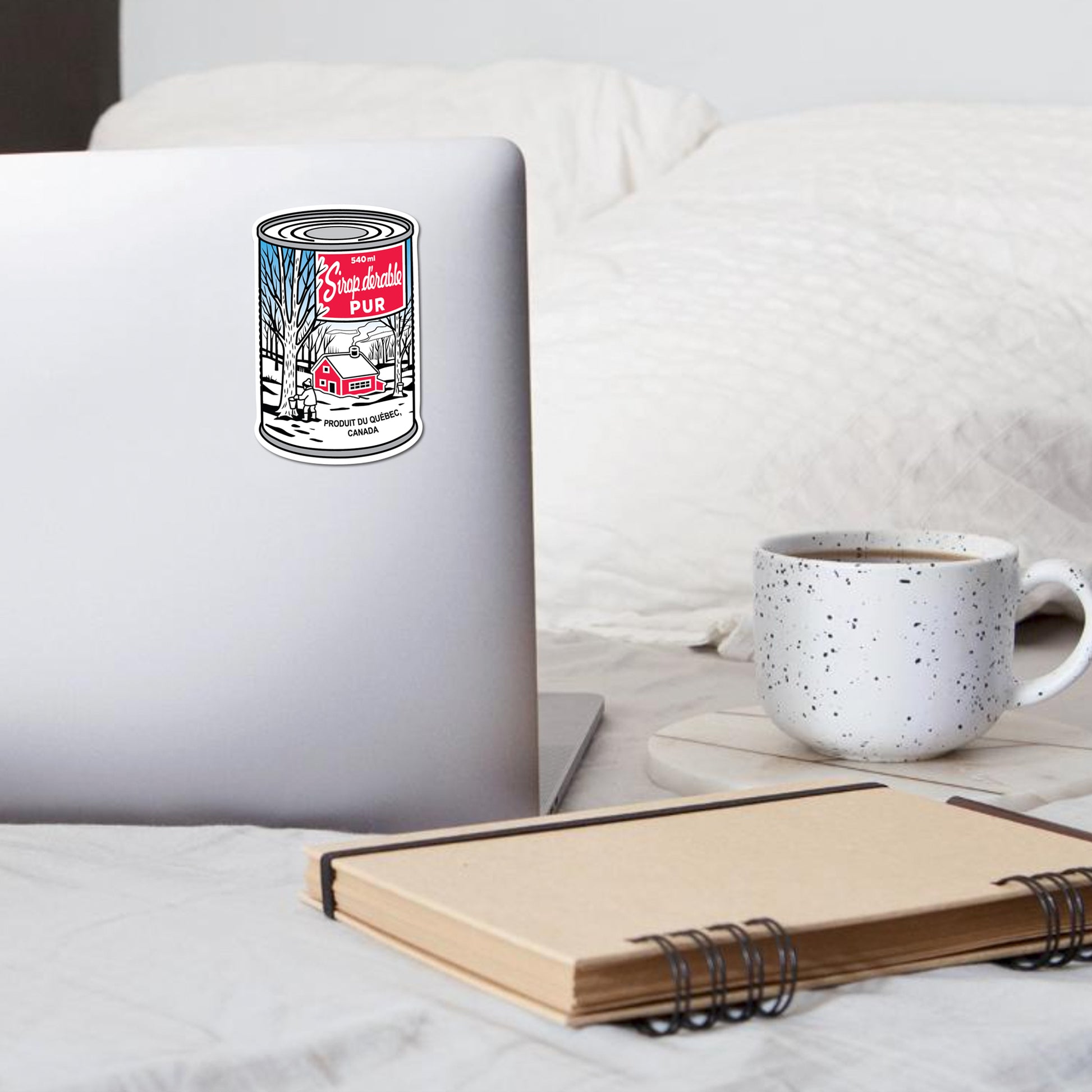 Laptop with a maple syrup can sticker on a bed next to a mug and notebook