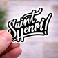 Hand holding a sticker with 'Saint Henri!' text against a blurred background