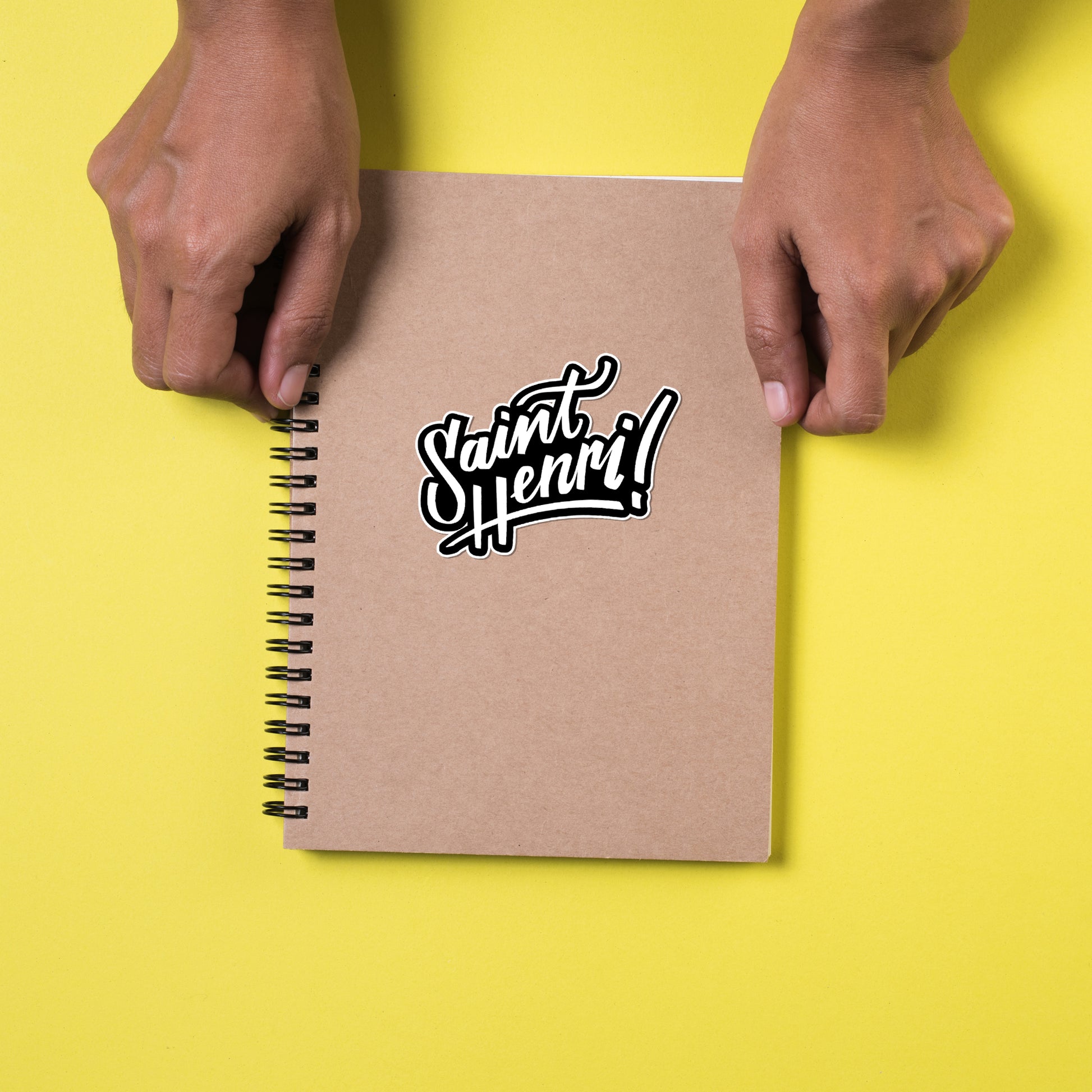 Notebook with 'Saint Henri' design held by hands on a yellow background