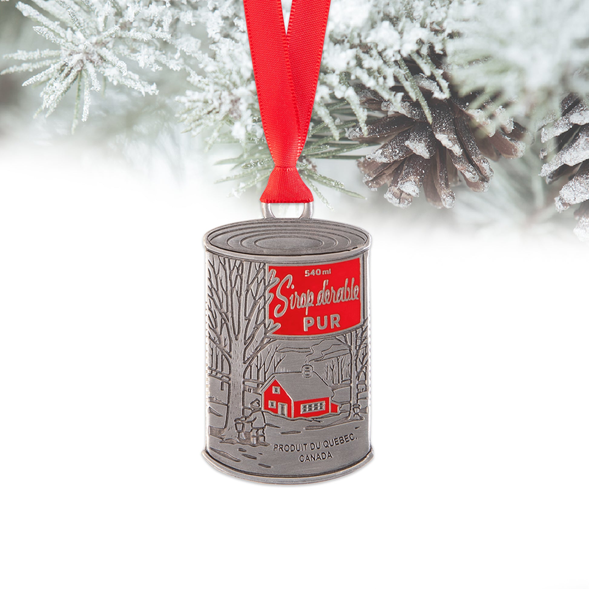 Decorative ornament shaped like a vintage maple syrop can with a red label, hanging from a red ribbon against a snowy tree background.