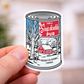 Sticker of a maple syrup can with a snowy scene and text, held by a hand against a blurred background.